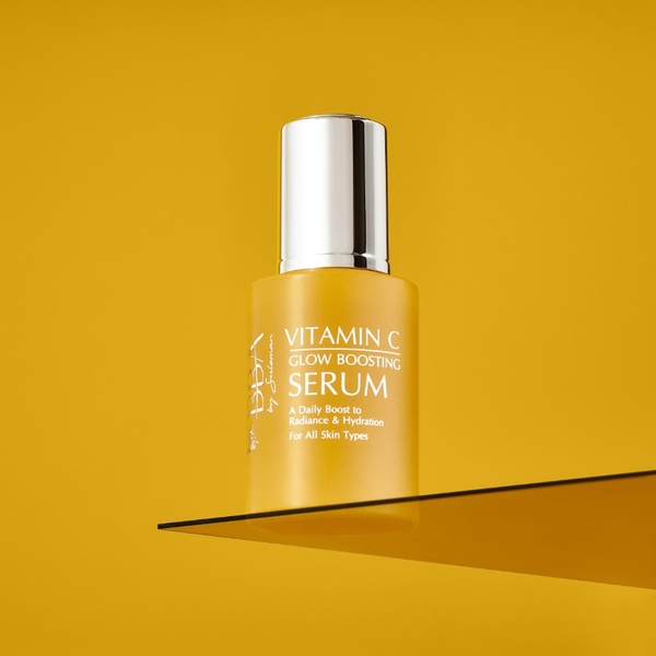 A clean, studio-lit hero shot of a Vitamin C glow boosting serum bottle perched on a glass shelf against a solid, vibrant yellow backdrop.